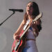 A woman with long dark hair stands on stage holding a red guitar and singing into a microphone on a stand. She is wearing a white crop top with blue stars on it and hot pants.