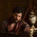 Kit Harington as Henry Muck, wearing a silk dressing gown, leaning over a grand piano