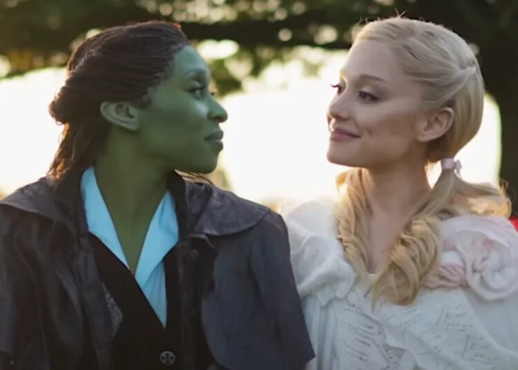 Cynthia Erivo's Elphaba and Ariana Grande's Glinda looking at each other smiling on the set of Wicked: For Good with poppy fields behind them.