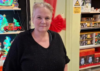 A woman with glasses on the top of her head and wearing a black top looks at the camera and smiles. She is standing among Christmas themed products in a shop.