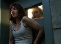 A woman looks scared while a man observes her through a small window in a bunker.