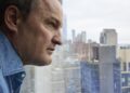 A close-up photo of a man looking out a window with the New York City skyline in the background