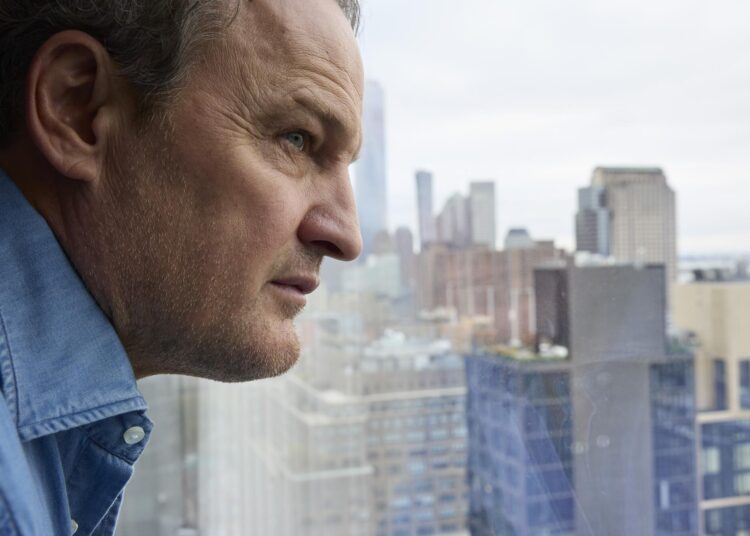 A close-up photo of a man looking out a window with the New York City skyline in the background