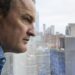 A close-up photo of a man looking out a window with the New York City skyline in the background
