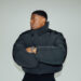 UK rapper Jay1 in a black jacket, standing against a wall with arms crossed.