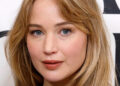 Jennifer Lawrence with long hair, wearing a simple black top, posing in front of a patterned backdrop