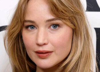 Jennifer Lawrence with long hair, wearing a simple black top, posing in front of a patterned backdrop
