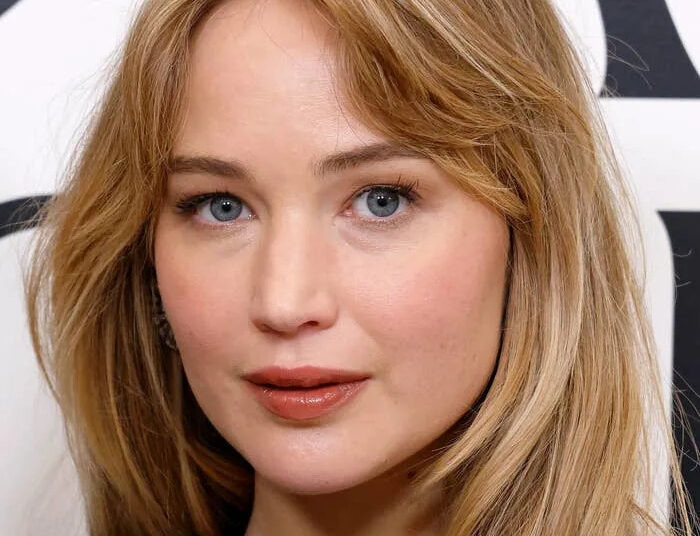 Jennifer Lawrence with long hair, wearing a simple black top, posing in front of a patterned backdrop