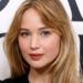 Jennifer Lawrence with long hair, wearing a simple black top, posing in front of a patterned backdrop