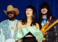 All three members of Khruangbin standing in front of a blue curtain, wearing eclectic, colorful clothing.