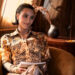 Kim Kardashian seated in a room, wearing a printed button-up shirt and matching pants, holding papers, and smiling