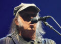 Neil Young sings into a microphone. He has long grey hair and sideburns. he is wearing a chequered navy jacket and a tan cap.