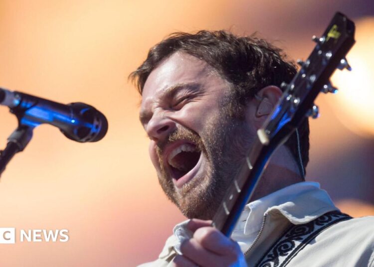 Kings of Leon lead vocalist Caleb Followill closes his eyes and sings into a microphone. He is wearing a light-coloured shirt and also playing a guitar.
