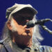 Neil Young sings into a microphone. He has long grey hair and sideburns. he is wearing a chequered navy jacket and a tan cap.
