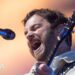 Kings of Leon lead vocalist Caleb Followill closes his eyes and sings into a microphone. He is wearing a light-coloured shirt and also playing a guitar.