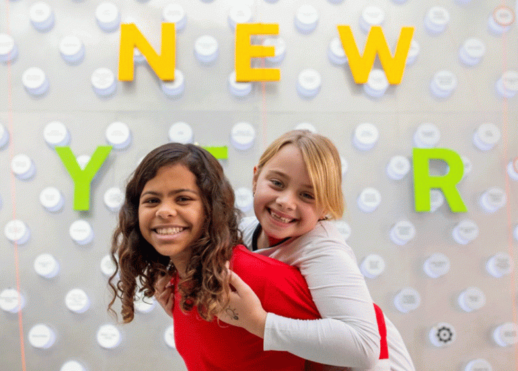 Two kids play piggy-back in front of a Happy New Year sign. (Courtesy photo)