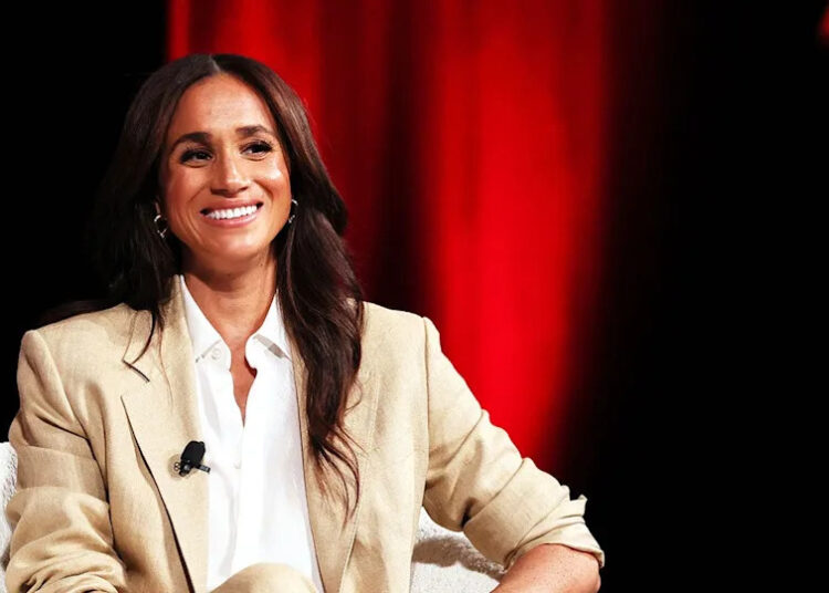 A close-up of Meghan Markle in a beige and white blouse smiling from a stage.