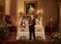 Elizabeth Hurley sat upright in a white coffin while Rob Rinder is stood in front of it, smiling ahead holding a red folder.