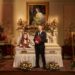 Elizabeth Hurley sat upright in a white coffin while Rob Rinder is stood in front of it, smiling ahead holding a red folder.