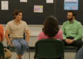 Two Latino men are seated on green chairs in a room in front of a large black board with four papers and a posted on it. The man on the left is wearing a beige short sleeved shirt and jeans. The man on the right has glasses and watch on his left hand. He is wearing a green long sleeved shirt and black pants. They are looking at each other in mid-conversation and are facing four people who are seated with their backs to the camera.
