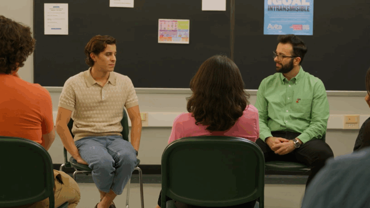 Two Latino men are seated on green chairs in a room in front of a large black board with four papers and a posted on it. The man on the left is wearing a beige short sleeved shirt and jeans. The man on the right has glasses and watch on his left hand. He is wearing a green long sleeved shirt and black pants. They are looking at each other in mid-conversation and are facing four people who are seated with their backs to the camera.