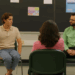 Two Latino men are seated on green chairs in a room in front of a large black board with four papers and a posted on it. The man on the left is wearing a beige short sleeved shirt and jeans. The man on the right has glasses and watch on his left hand. He is wearing a green long sleeved shirt and black pants. They are looking at each other in mid-conversation and are facing four people who are seated with their backs to the camera.
