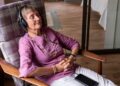 Senior woman relaxing in chair with eyes closed, hands folded listening to music with headphones on and smartphone on lap.