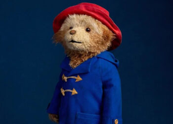 The musical's Paddington bear stands wide-eyed in front of a dark blue background. He has a toy-like quality, wearing his famous blue duffle coat and large red hat.
