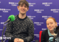 A 20-year-old man and 13-year-old girl sit in front of microphones in a radio studio, smiling at the camera.