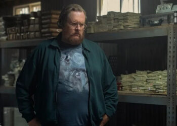 Steve Agee as John Economos looks serious in front of a lot of money, wearing a glorious three raccoon moon shirt