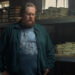 Steve Agee as John Economos looks serious in front of a lot of money, wearing a glorious three raccoon moon shirt