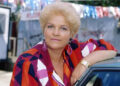 Pam St Clement as Pat Butcher in 1992 in a bright red, purple and orange blouse with large purple earrings, and leaning on a car roof