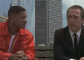 Two individuals sit on a bench; one wears a red jacket with a white shirt, and the other in a suit, engaged in conversation against a city backdrop