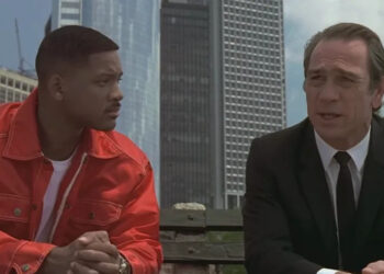 Two individuals sit on a bench; one wears a red jacket with a white shirt, and the other in a suit, engaged in conversation against a city backdrop