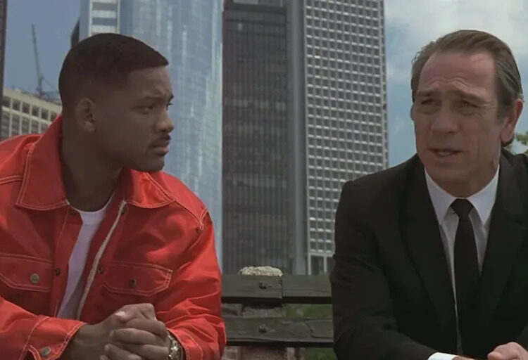 Two individuals sit on a bench; one wears a red jacket with a white shirt, and the other in a suit, engaged in conversation against a city backdrop