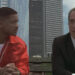 Two individuals sit on a bench; one wears a red jacket with a white shirt, and the other in a suit, engaged in conversation against a city backdrop