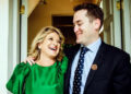 Sam wearing a suit with his arm around his mother in an emerald dress looking at each other and laughing.