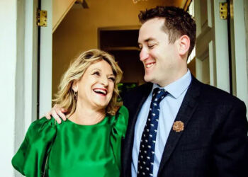 Sam wearing a suit with his arm around his mother in an emerald dress looking at each other and laughing.