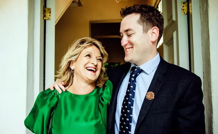 Sam wearing a suit with his arm around his mother in an emerald dress looking at each other and laughing.