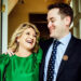 Sam wearing a suit with his arm around his mother in an emerald dress looking at each other and laughing.