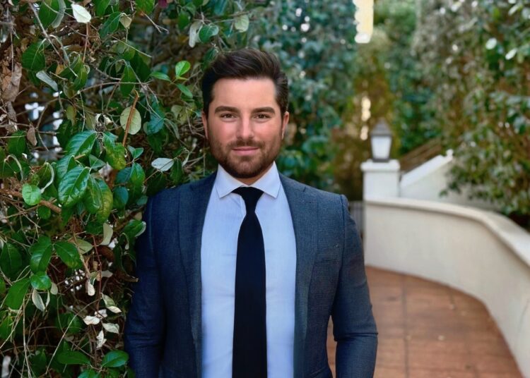 Kyle Boulia wearing a blue suit and tie posing in front of a bush and a stone sidewalk