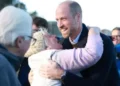 PA Media A older woman with white hair, wearing a pink jumper, gilet and a woolly hat, embraces Prince William as she looks up at him. Prince William smiles as people around them watch.