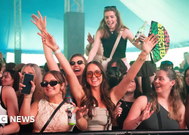 A crowd of people in a big festival tent with hands up and smiling into the camera. Someone is up on their friends shoulders and one girl is holding a "defected records" fan.