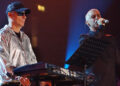 The Pet Shop Boys on stage in 2022, Chris Lowe, on the left is wearing a silver jacket, cap and glasses as he stands next to a keyboard. Neil Tennant stands close to him holding a microphone to his mouth.