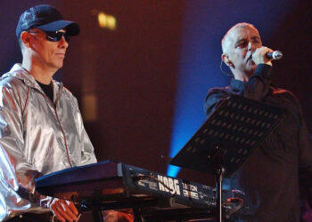 The Pet Shop Boys on stage in 2022, Chris Lowe, on the left is wearing a silver jacket, cap and glasses as he stands next to a keyboard. Neil Tennant stands close to him holding a microphone to his mouth.