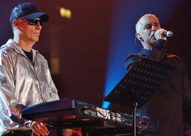 The Pet Shop Boys on stage in 2022, Chris Lowe, on the left is wearing a silver jacket, cap and glasses as he stands next to a keyboard. Neil Tennant stands close to him holding a microphone to his mouth.
