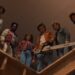 Six teenagers standing together on a ledge within a building. The teens are all looking down at something in confusion.