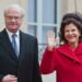 The King and Queen of Sweden stand side by side. The Queen is in red and is waving the king is standing with his arms by his side.