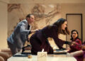 A lively boardroom scene with four people, including one standing on a table. They're engaged in an animated discussion