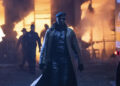 Lee Pace as Evan McCone wearing a mask and trench coat standing in front of a burning building in The Running Man